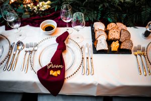 Elegant table setting at Dodmoor House, Northamptonshire's best wedding venue, featuring a maroon napkin labeled "Groom," cutlery, wine glasses, bread slices with butter, and a small dish of dip on a pristine white tablecloth. Perfect for capturing by a Dodmoor House Wedding Photographer.