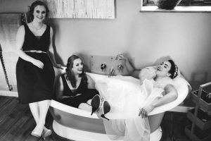 At Dodmoor House, Northamptonshire's best wedding venue, three women find humor in a bathroom moment. One sits on the tub edge, another inside it, while a bride reclines in laughter within the bathtub—all caught candidly by a Dodmoor House Wedding Photographer.
