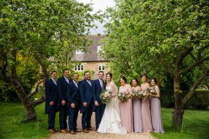 A wedding party poses on a tree-lined path, the bride and groom at the center, flanked by bridesmaids in mauve and groomsmen in navy, capturing the essence of Dodmoor House Wedding Photography at this picturesque Northamptonshire venue.