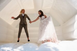 In their formal attire, two people joyfully jump hand in hand on a white inflatable bounce house, capturing a delightful moment perfect for Dodmoor House Wedding Photography.