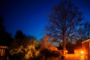 As dusk falls over the backyard, string lights adorn the trees and bushes, casting a warm glow reminiscent of a Dodmoor House wedding venue. The deep blue sky cradles a faint celestial body, while the patio with chairs invites you to sit back and take in this serene scene.
