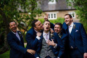 Five men in suits having fun outdoors; one man is being playfully licked and held in a humorous pose. Captured at Dodmoor House, they are all laughing, wearing matching dark blue suits with light purple ties—perfect for Dodmoor House Wedding Photography.