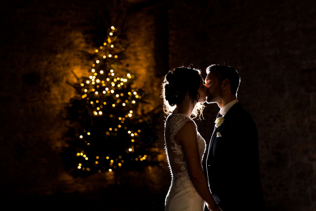 Couple kissing in front of a glowing Christmas tree, with dim lighting creating a romantic atmosphere.