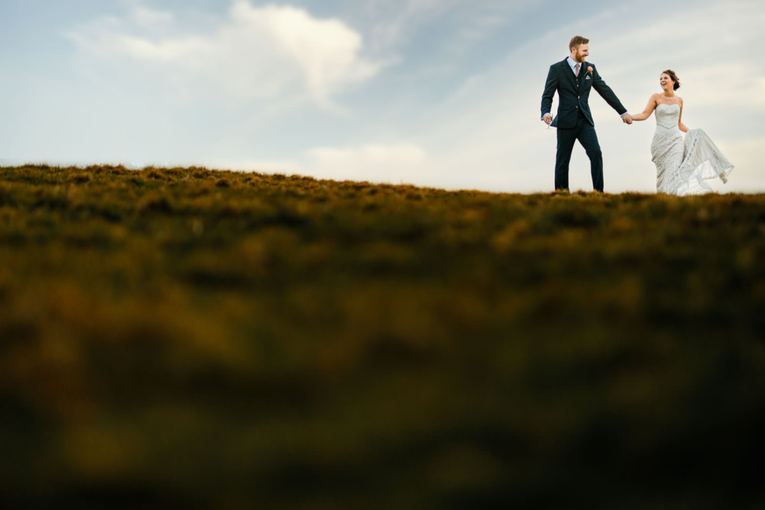Bride and groom holding hands, walking on a grassy hill under a cloudy sky, capturing the enchanting essence of winter wedding photography.