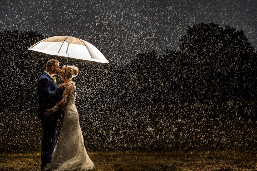 A couple in wedding attire stands under an umbrella, embracing in the rain's embrace at their winter wedding, surrounded by darkness.