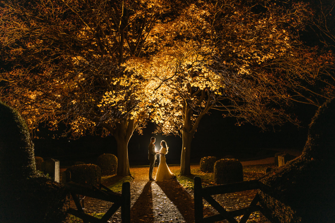 A couple stands under a tree with illuminated golden leaves at night, creating a dramatic and romantic scene perfect for winter wedding photography.