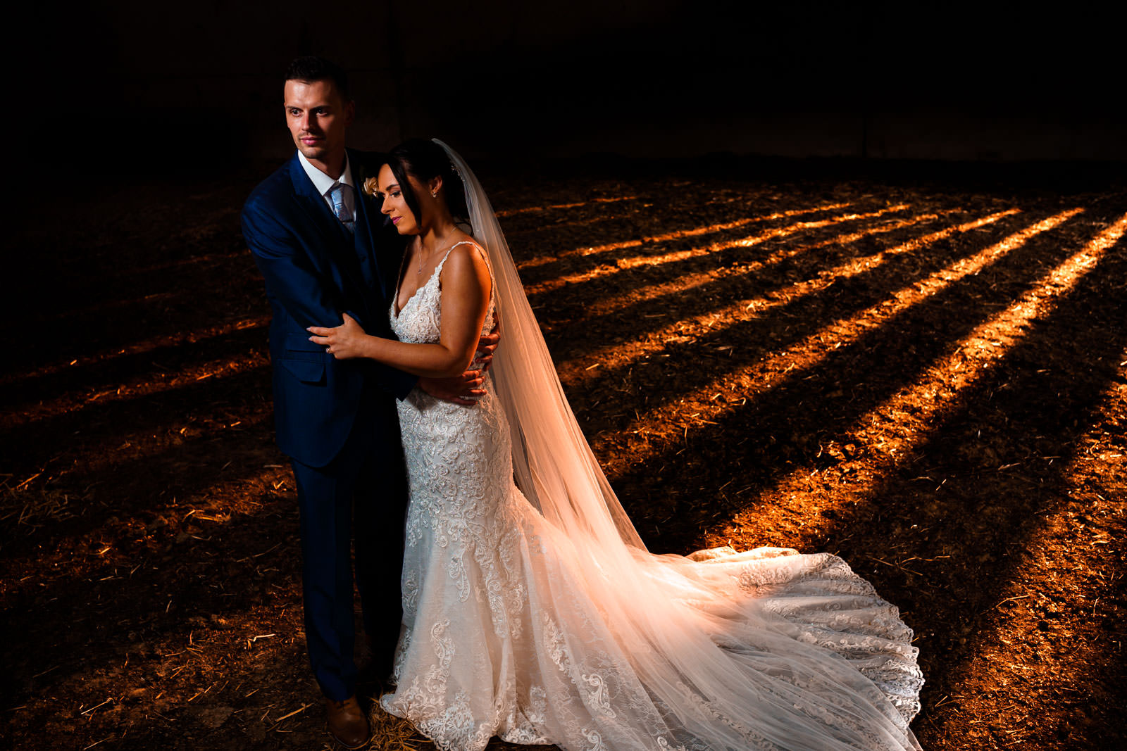 Bride and groom stand together in dim lighting with sunlit stripes on the ground. Bride wears a lace gown with a long train, and the groom is in a dark suit.
