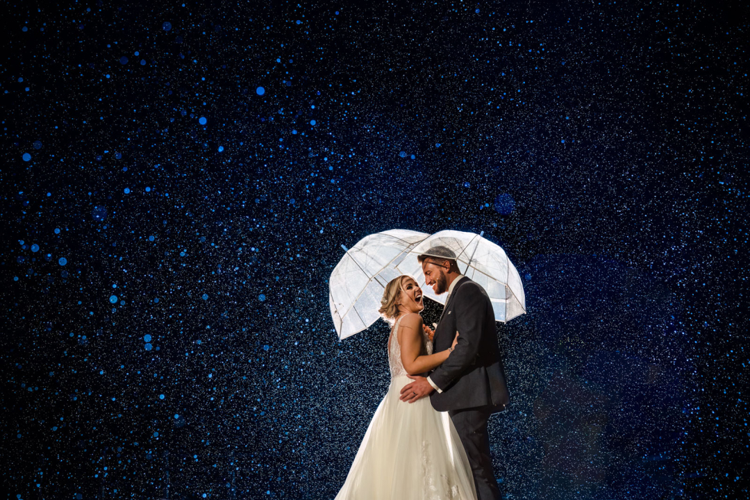 A couple in wedding attire stands under a transparent umbrella, captured by a skilled winter wedding photographer, surrounded by the enchanting backdrop of falling rain and a starry night sky.
