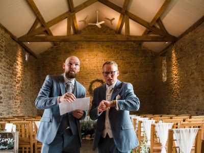 second photographer photos Two men in suits stand in a wood-beamed venue; one holds a paper, the other checks his watch, contemplating the benefits of a second photographer. Rows of chairs are arranged neatly for an event.