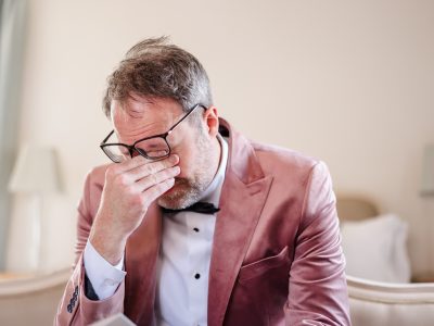 second photographer shots 11 A man in a pink suit and glasses sits indoors, pinching the bridge of his nose with a pained expression, perhaps contemplating the benefits of a second photographer for groom prep photography.