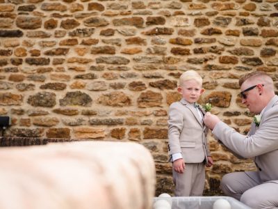 second photographer shots 17 A man in sunglasses adjusts the boutonniere of a young boy in a suit, standing against a brick wall. Capturing such candid moments highlights the benefits of two wedding photographers, as one focuses on groom prep photography while another captures vibrant details.