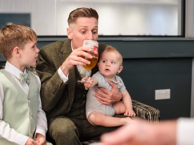 second photographer shots 18 A man in a suit sips a beer while holding a baby, sitting next to a young boy in formal attire—capturing one of those unexpected moments that make you wonder if you should hire two photographers for flawless groom prep photography.