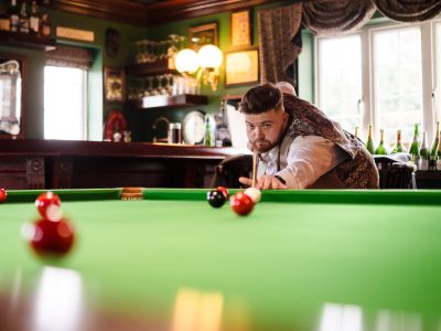 second photographer shots 6 A person focuses intently, aiming at a billiard ball on the green pool table in a dimly lit room. Wooden furnishings and bottled drinks line the shelves like a perfect shot captured by talented husband and wife photographers.