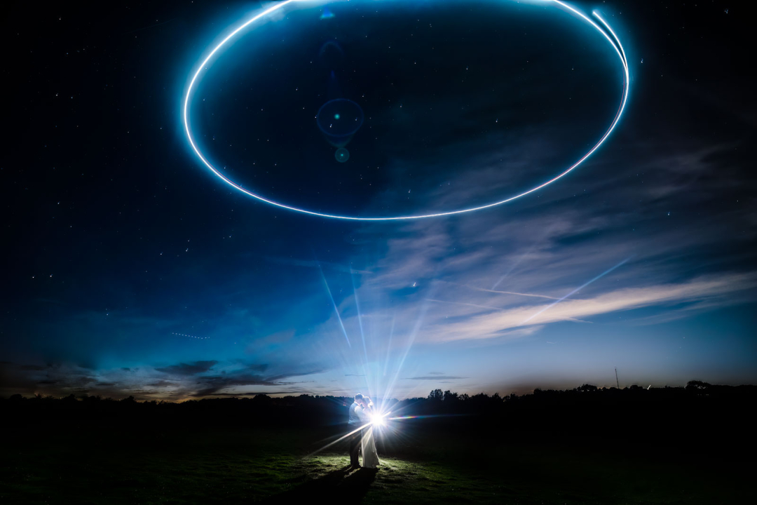 A couple embraces under a night sky with a glowing blue ring of light above them, reminiscent of enchanting winter wedding photography ideas, set against fading twilight and scattered clouds.