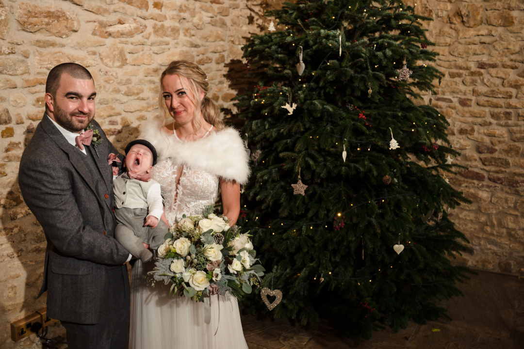 A man and woman pose with a baby in formal attire beside a decorated Christmas tree.