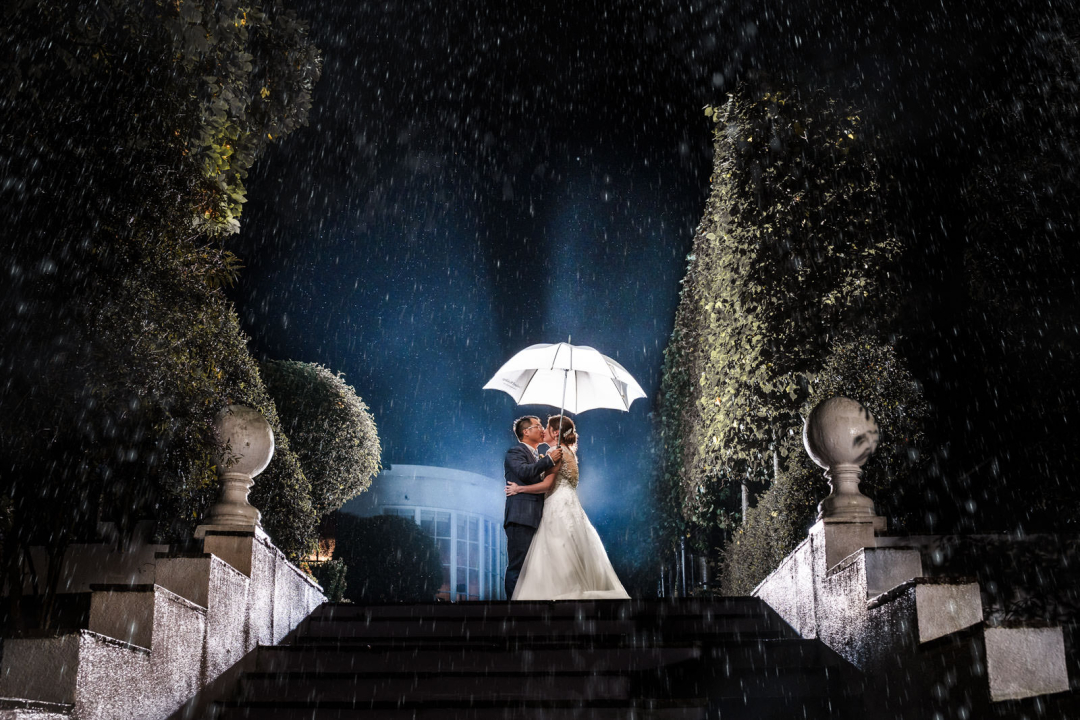 A couple in wedding attire stands under a white umbrella on stone steps, surrounded by lush greenery and a gentle rain, with a bright light behind them. This enchanting scene is perfect for winter wedding photography ideas that capture the romance of the season.