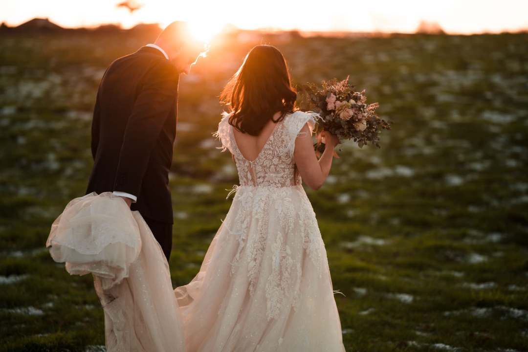 Bride in a white dress and groom in a suit walk hand in hand across a snowy field at sunset, capturing the magic of winter wedding photography.
