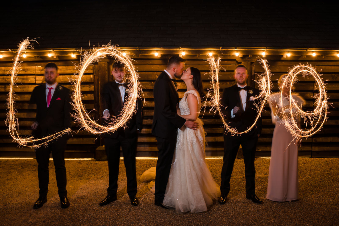 Five people holding sparklers forming "LOVE" with a couple kissing in the center, dressed in wedding attire, with string lights in the background.