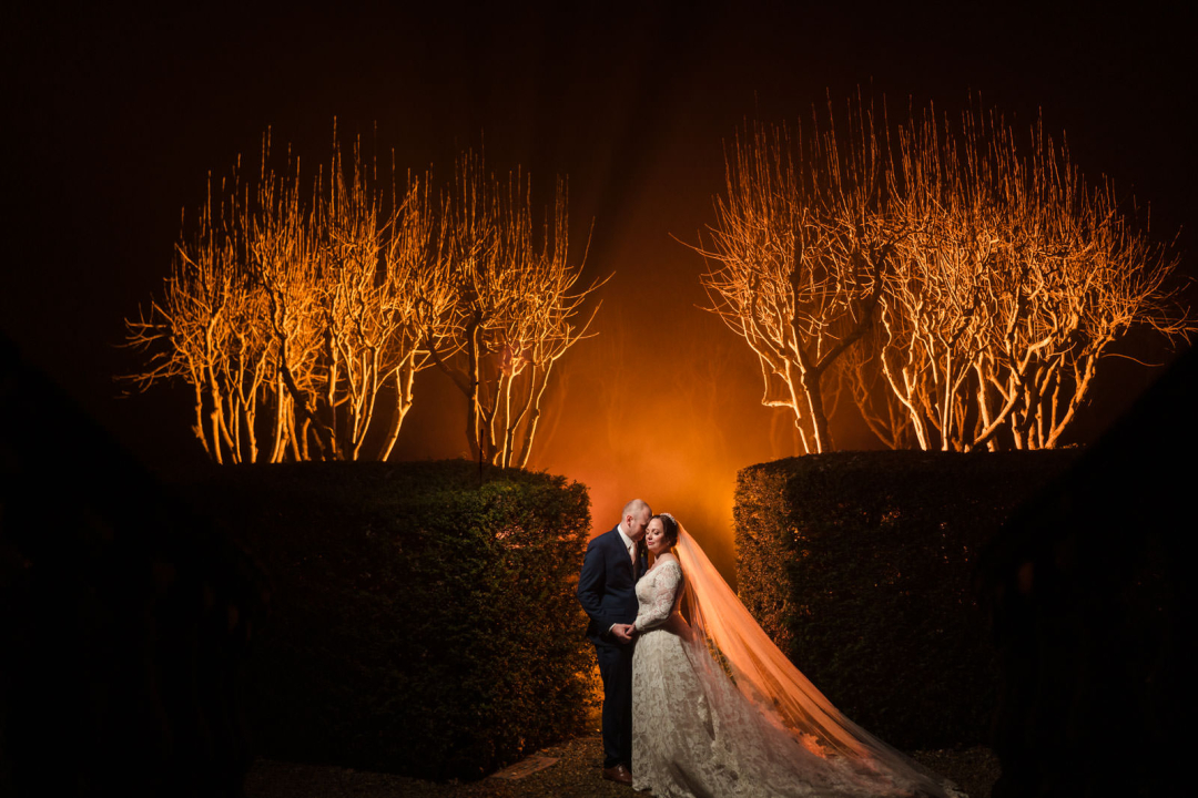 Bride and groom stand embracing at night, with illuminated bare trees and hedges in the background, creating a dramatic, warm glow. Perfect inspiration for those seeking winter wedding photography ideas.