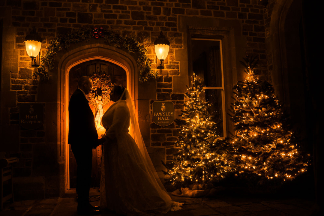 A couple in wedding attire holds hands at night in front of an illuminated doorway, embraced by festively decorated Christmas trees and outdoor lamps—capturing the essence of magical winter wedding photography ideas.