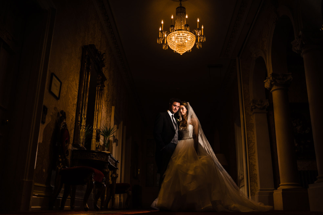 Bride and groom stand together in an elegantly lit hallway under a chandelier, capturing the essence of winter wedding photography with ornate decor and arches in the background.