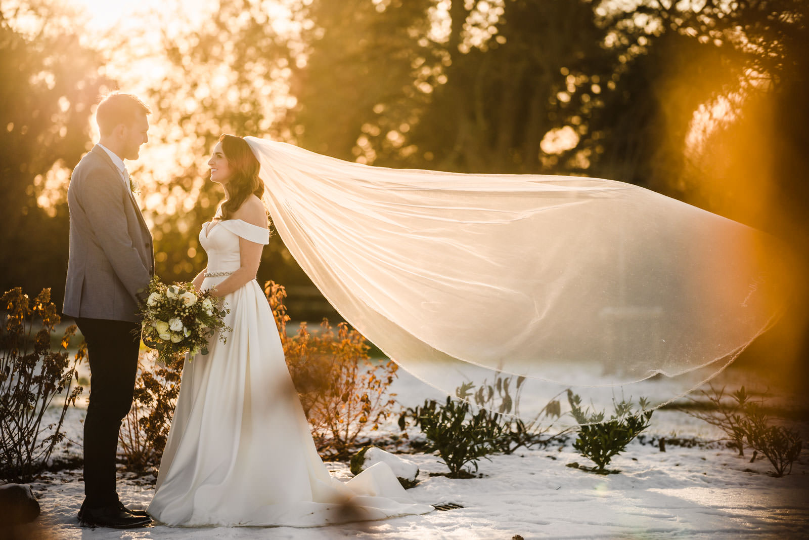 Bride and groom standing outdoors, the bride's veil flowing in the wind, surrounded by snow-covered ground and sunlight filtering through trees—an enchanting scene perfect for winter wedding photography ideas.