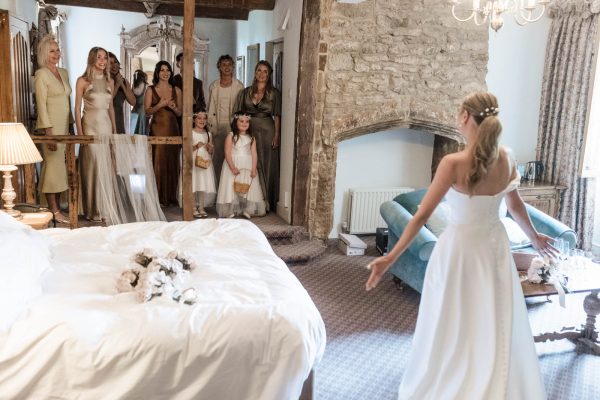 A bride in a white dress faces her bridesmaids and flower girls in a rustic bedroom with stone walls and wooden beams, beautifully captured by a Fawsley Hall photographer.