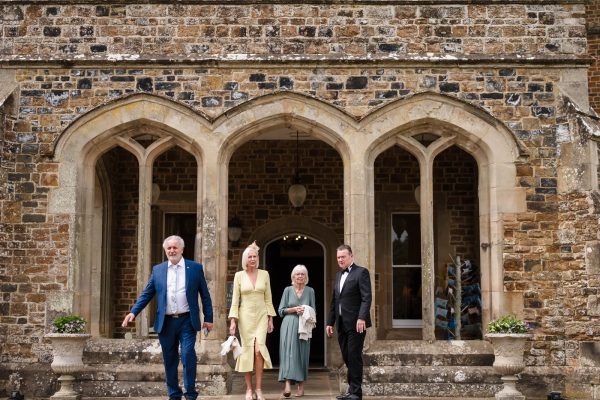 Four adults dressed formally stand in front of the historic stone building, with arched doorways and two potted plants flanking the entrance—an elegant moment captured by a Fawsley Hall photographer.