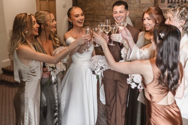 A bride, groom, and wedding party stand together indoors at Fawsley Hall, raising glasses in a joyful toast and smiling—a perfect moment captured by a skilled Fawsley Hall photographer.