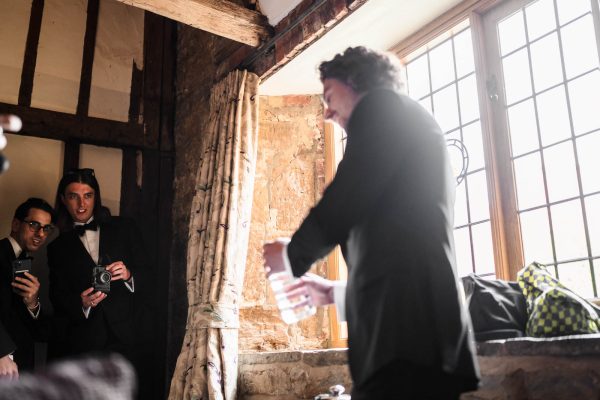 Three men in suits stand indoors near a window at Fawsley Hall; two hold cameras and watch another man, who is holding a glass and is partially out of focus, capturing the essence of Fawsley Hall photography.