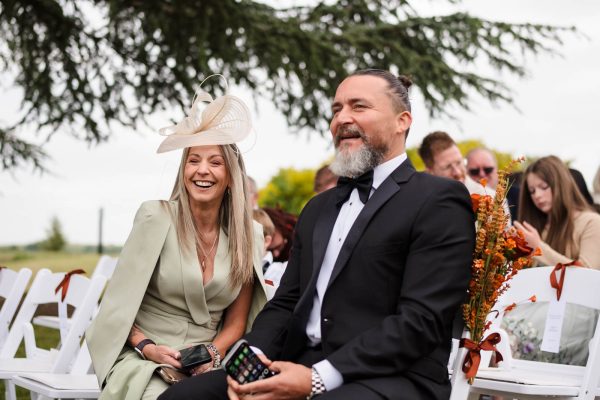 A woman in a light green dress and hat sits next to a man in a black suit and bow tie at an outdoor event, beautifully captured by Fawsley Hall photography, with guests gathered in the background.