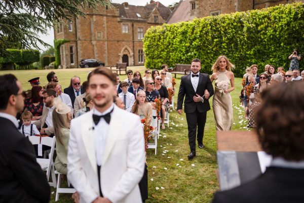 A bridesmaid and groomsman walk down an outdoor wedding aisle lined with seated guests, as the grandeur of Fawsley Hall sets a stunning backdrop—perfect for timeless Fawsley Hall photography.