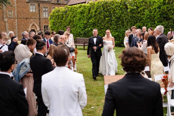 A bride in a white gown walks down an outdoor aisle at Fawsley Hall with an older man, surrounded by seated and standing wedding guests.