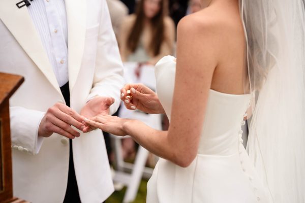 A bride and groom exchange rings during an outdoor wedding ceremony at Fawsley Hall. The bride wears a strapless white dress and veil; the groom wears a white jacket, beautifully captured by Fawsley Hall photography.