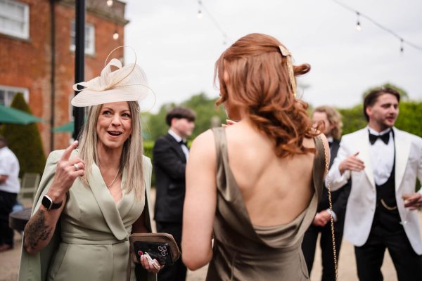 Two women in formal attire converse at an outdoor event at Fawsley Hall, with other guests in suits visible in the background, perfectly capturing a candid moment of elegance by a Fawsley Hall photographer.