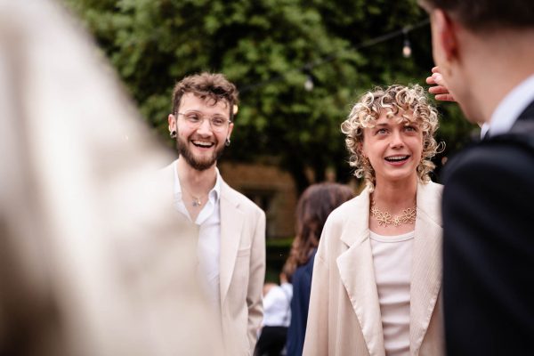 Two people in light-colored suits smile and chat with others outdoors at Fawsley Hall, surrounded by green trees and string lights—perfectly captured by a skilled Fawsley Hall photographer.