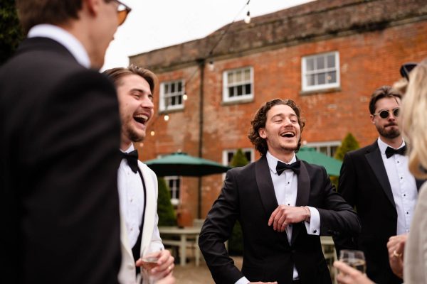 A group of men in tuxedos stand outside a brick building, laughing and talking together at what appears to be a formal event, beautifully captured by Fawsley Hall photography.