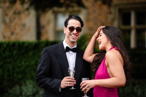 A man in a tuxedo and sunglasses and a woman in a pink dress laugh together outdoors while holding glasses, beautifully captured by Fawsley Hall photographer.