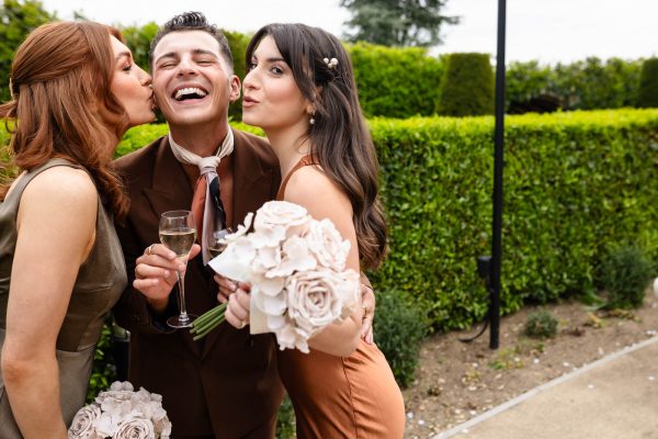 Two women in formal attire kiss a man on the cheek while he holds a glass of wine and smiles, standing outdoors near green hedges. Both women hold bouquets of pale flowers—captured perfectly by a Fawsley Hall photographer.