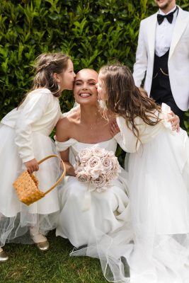 A bride smiles as two young girls in white dresses kiss her cheeks; she holds a bouquet of roses, and a man in a white suit stands nearby—a magical moment captured by a Fawsley Hall photographer.