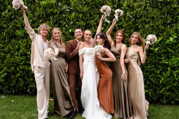 A newlywed couple poses with five women holding bouquets, all standing in front of a green leafy backdrop in coordinated earth-toned outfits, perfectly captured by a Fawsley Hall photographer.