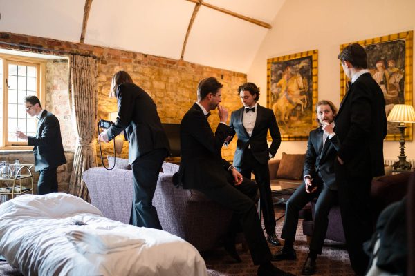 Six men in suits gather in a stylish, rustic room at Fawsley Hall; some are talking, one is using a camera—capturing moments for Fawsley Hall photography—and another pours a drink near the window.