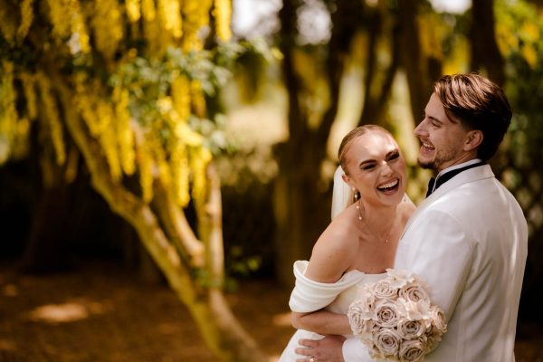 A bride holding a bouquet of roses and a groom in a white suit jacket smile and embrace outdoors in front of yellow flowering trees, captured beautifully by a Fawsley Hall photographer.