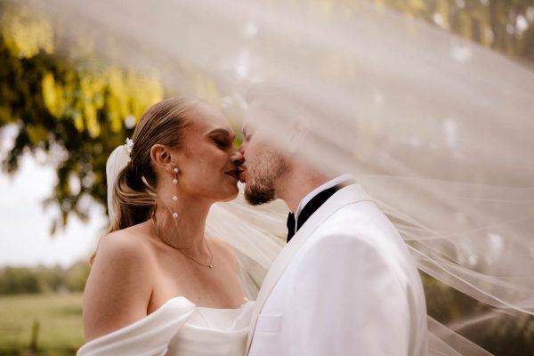 A bride and groom in white attire share a kiss outdoors at Fawsley Hall, partially covered by the bride’s veil with lush greenery in the background, perfectly captured by a Fawsley Hall photographer.