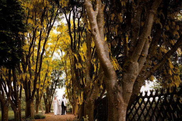 A couple dressed in formal attire walks along a tree-lined path with yellow blossoms at Fawsley Hall, surrounded by fencing and greenery—an elegant moment captured by a skilled Fawsley Hall photographer.