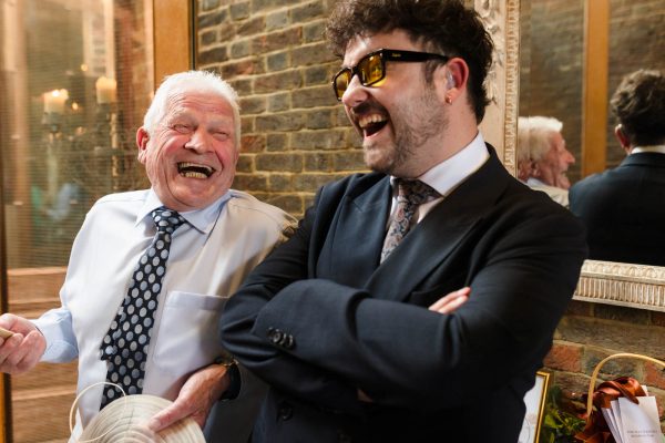 An older man in a dress shirt and tie laughs with a younger man in a suit and sunglasses; both appear to be enjoying a light-hearted moment indoors, perfectly captured by Fawsley Hall photography.