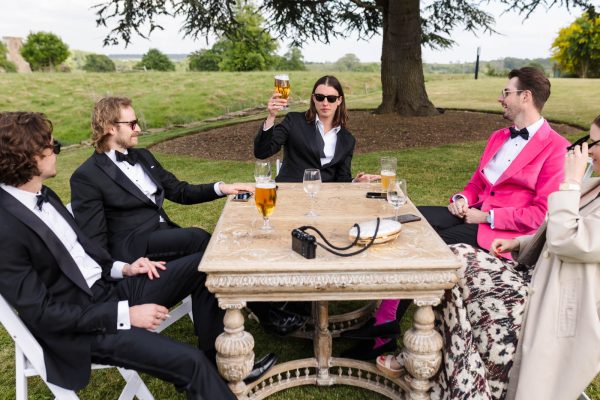 Four men in formal wear and one person in a coat sit around a wooden outdoor table with drinks, a camera, and a phone on the grassy lawn of Fawsley Hall, making for the perfect setting for Fawsley Hall photography.