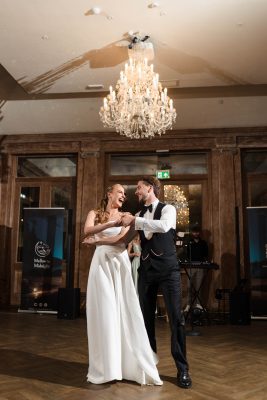 A bride and groom dance together, smiling, in a ballroom at Fawsley Hall with a chandelier overhead and a musician performing in the background—captured by a skilled Fawsley Hall photographer.