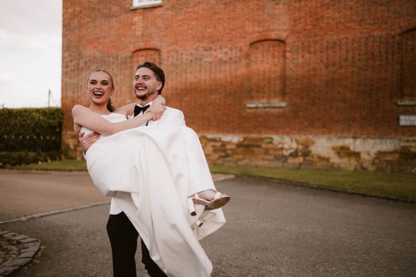 A groom in a tuxedo carries his laughing bride in a white dress outside near a brick building on a paved path, captured beautifully by a Fawsley Hall photographer.