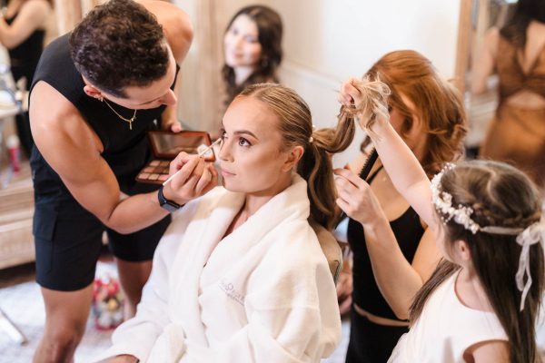 A woman in a white robe gets her makeup done by a man while another person styles her hair and a young girl holds a hair strand, beautifully captured by a Fawsley Hall photographer.